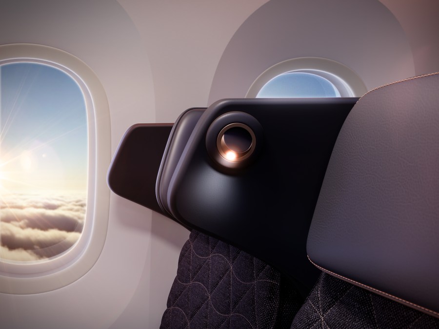 Close-up of an airplane seat with a view of clouds through the window, featuring a sleek headrest design and a reading light.